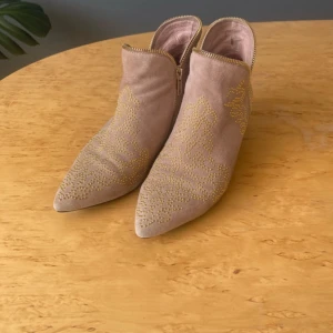 Beige mocka boots med gulddetaljer - Snygga boots i beige mocka med spetsig tå och låg klack. Skorna har dekorativa guldfärgade nitar i mönster på ovandelen och dragkedja längs sidan. Perfekta för dig som vill sticka ut med en cool och trendig look.
