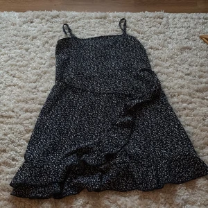 Blå blommig kortklänning med volang - Supersöt blå kortklänning med vita småblommor och omlott volangkant nertill. Väldigt skönt och luftigt material perfekt till en varm sommardag! 