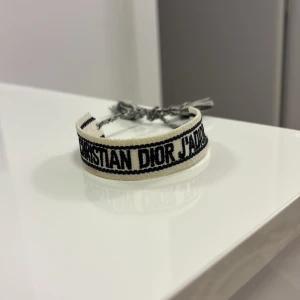 Christian Dior J'adior armband - Snyggt armband från Christian Dior i vit och svart textil med broderad text 'J'adior'. Justerbar med flätade band och tofsar för en cool och personlig look. Perfekt accessoar för att lyfta din outfit med en lyxig touch.