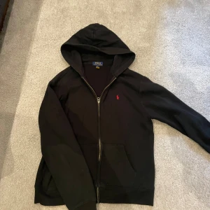 Svart zip up hoodie från Polo Ralph Lauren storlek S - Knappt använd Svart zip up hoodie från Polo Ralph Lauren i storlek S. Klassisk design med dragkedja, huva och två fickor framtill. Ikoniska röda loggan broderad på bröstet. Mjuk bomullskänsla och perfekt för chill dagar. (Nypris 1200kr)