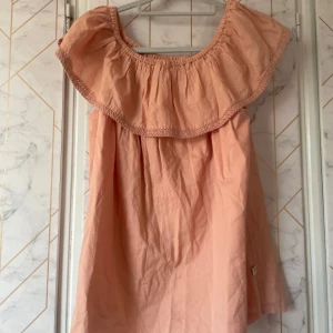 Rosa blus från Pomp de Lux - Supersöt rosa blus från Pomp de Lux med bred volang runt axlarna och smockad halsringning. Luftig modell och är ärmlös. I storlek 158/164