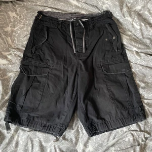 Svarta cargoshorts med snörning - Svarta cargoshorts med flera stora fickor, snörning i midjan och justerbara spännen i sidorna. Shortsens material känns slitstarkt och de har en loose passform. Perfekta för chill dagar och eller när du vill ha en streetwear stil.  