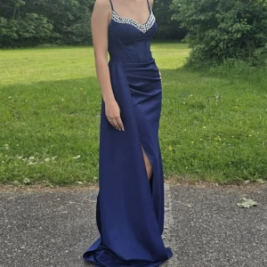 Marinblå balklänning med strass - Säljer en marinblå långklänning med smala axelband och snygg slits framtill. Klänningen har en figurnära passform och är dekorerad med glittrande strass längs urringningen. Materialet är slätt och känns lyxigt, perfekt för bal eller fest.💕 Pris kan diskuteras.