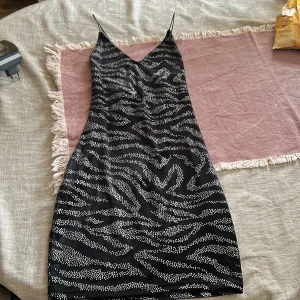 Zebramönstrad kort klänning H&M Divided - Snygg svart kort klänning från H&M Divided med tunna axelband och v-ringning. Klänningen har ett zebramönster i silverprickar som ger en glittrig effekt. Perfekt för dig som vill sticka ut med en trendig look.