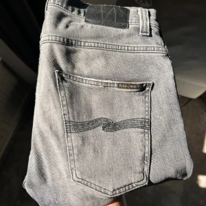 Grå jeans från Nudie Jeans Co - Säljer ett par grå jeans från Nudie Jeans Co med klassisk femficksdesign och svarta sömmar på bakfickan. Jeansen har en rak passform och är tillverkade i denim. Perfekta för dig som gillar en stilren look. Jeansen har några snygga slitningar på sig också.
