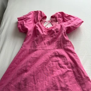 Rosa klänning med puffärmar och knyt i ryggen - Säljer en söt rosa klänning med puffiga kortärmar och v-ringning. Klänningen har knytband i ryggen och är sydd i ett lätt, vävt material som känns luftigt. 