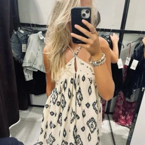 Zara sommarklänning - Säljer en luftig vit klänning med svarta grafiska mönster och tunna axelband. Klänningen har en liten v-ringning med knappdetalj framtill och är ärmlös. Perfekt för varma dagar!
