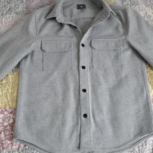 Ljusgrå overshirt från H&M - Snygg ljusgrå overshirt från H&M med klassisk krage, svarta knappar och två stora bröstfickor med lock. Skjortan har lång ärm och en rak, något loose passform. Perfekt att slänga över en t-shirt för en chill look. Priset kan diskuteras.