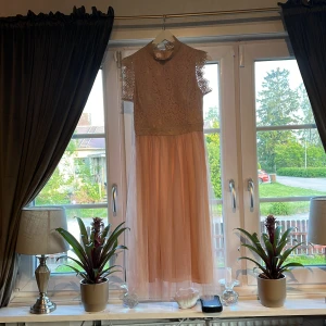 Rosa långklänning med spets och tyll - Säljer en elegant långklänning i rosafärgad  med överdel i spets och ärmlös design. Kjolen är i mjuk tyll som ger ett luftigt intryck. Perfekt för bal eller fest. Hög halsringning och fina spetsdetaljer på axlarna.