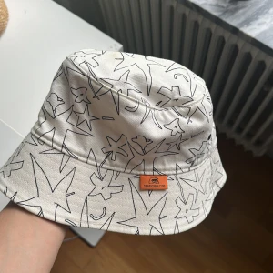 Vit bucket hat från Zara med print - Vit bucket hat från Zara med svart grafiskt stjärn- och blomprint designat av Sebastian Curi. Hatten har en klassisk form och är tillverkad i bomull, perfekt för att lyfta din sommarstil med en cool och unik vibe.