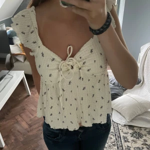 Blommig volangblus med knyt - Supersöt blus i ljusbeige med små blommor, volangärmar och knyt framtill. Luftig och flowy modell med lätt rynkat tyg, perfekt för varma dagar. Sitter snyggt och ger en romantisk vibe. Aldrig använd!!