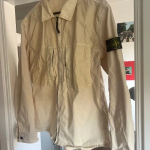 Beige overshirt från Stone Island - Snygg beige overshirt från Stone Island med klassisk patch på ärmen. Skjortan har två stora bröstfickor, dragkedja framtill och lång ärm. Perfekt för lager-på-lager och streetwear-vibe.