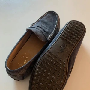 Mörkblå loafers från Polo Ralph Lauren - Snygga mörkblå loafers från Polo Ralph Lauren i textil med klassisk pennyloafers-design. Skorna har dekorativa sömmar och gummisula med nitar för extra grepp. Perfekta för en stilren och avslappnad look.
