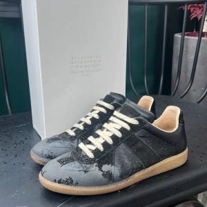 Maison Margiela GATS Paint Splatter - Maison Margiela sneakers i svart skinn med beige sula och vita snören. Klassisk låg modell med rund tå och platt sula. Lämnat in till skomakare för att sy på mocka vid insidan av hälen så de glider mindre. Kommer med originalkartong, dustbag och kvitto från Paul & Friends NK där jag köpte dem. 