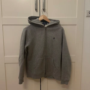 Grå hoodie från Polo Ralph Lauren - Klassisk grå hoodie från Polo Ralph Lauren med dragkedja och huva. Tröjan har två fickor framtill och en liten broderad logga på bröstet. Perfekt för en avslappnad stil. Tröjan är från kidsbrandstore och har storlek xl i barn storlek vilket betyder att den passsar upp till 178cm lång.