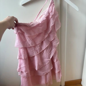 Rosa volangklänning med oneshoulder - Söt rosa kortklänning med flera volanglager och oneshoulder-design. Klänningen har tunna axelband och är tillverkad i ett lätt, luftigt material som ger en romantisk känsla. Perfekt för sommarens tillfällen när du vill sticka ut lite extra.