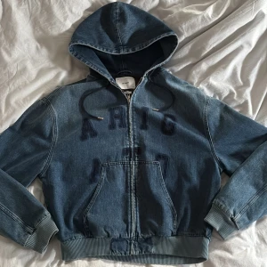 Jeansjacka med huva från Axel Arigato - Säljer en blå jeansjacka med huva från Axel Arigato. Jackan har dragkedja framtill, ribbade muddar och fickor på magen. Broderad text på bröstet och justerbara snören i huvan. Köpt på Axel Arigatos hemsida för ca 3000kr