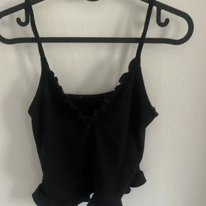 Svart croppad linnetopp med volang - Säljer en svart croppad linnetopp med smala axelband och volangdetalj längs urringningen. Toppen har en ribbad struktur och är perfekt för varma dagar.