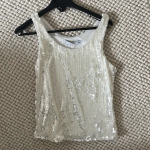 Vit paljettprydd linnetopp från Mango  - Säljer en snygg vit linnetopp från Mango som är täckt med glittrande paljetter. Perfekt för att sticka ut på festen! Toppen är ärmlös och har en rund halsringning.