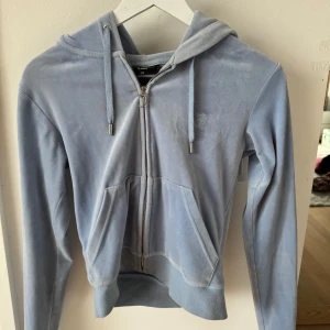 Ljusblå velour hoodie från Juicy Couture - Säljer en ljusblå hoodie i mjuk velour från Juicy Couture. Tröjan har dragkedja, huva med snören och två fickor framtill. Klassisk broderad JC-logga på bröstet.