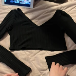 Svart ribbad croppad topp Chiquelle - Säljer en svart croppad topp från Chiquelle med lång ärm och djup v-ringning. Toppen är ribbad och har en tight passform som framhäver siluetten. Perfekt att styla med höga jeans eller kjol för en trendig look.