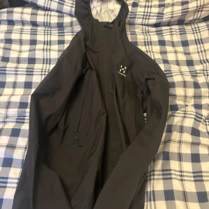 Svart vindjacka från Haglöfs med huva - Svart vindjacka från Haglöfs med huva och dragkedja framtill. Jackan har minimalistisk design, diskret logga på bröstet och praktiska fickor med dragkedja. Perfekt för blåsiga dagar och outdoor-äventyr.