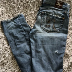 Blå jeans från Rock & Republic - Snygga blå jeans från Rock & Republic med slitningar och lätt tvättad look. Klassisk femficksmodell med låg midja och raka ben. Jeansen har detaljer som slitna kanter och en liten reva på benet för en cool, avslappnad stil.