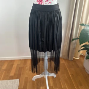 Svart plisserad kjol med mesh från Twist - Säljer en svart plisserad kjol från Twist med bred midja och transparent mesh nedtill. Kjolen har en snygg, flowy siluett och är perfekt för dig som vill sticka ut lite extra. Midilängd med cool layering-effekt.