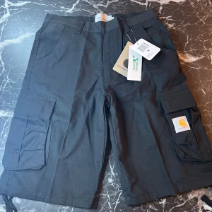 Svarta cargoshorts från Carhartt WIP - Säljer ett par svarta cargoshorts från Carhartt WIP med flera stora fickor på sidorna och klassisk Carhartt-logga. Shortsen har normal passform och är tillverkade i slitstark bomull. Perfekta för en avslappnad stil.
