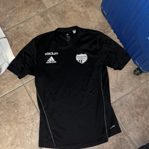 Svart Sollentuna FK fotbollströja Adidas - Svart fotbollströja från Adidas med Sollentuna FK-logga på bröstet. Tröjan har korta ärmar, rund hals och är tillverkad i lätt climalite-material som andas. Adidas och Stadium-loggor i vitt framtill. Perfekt för träning eller match.