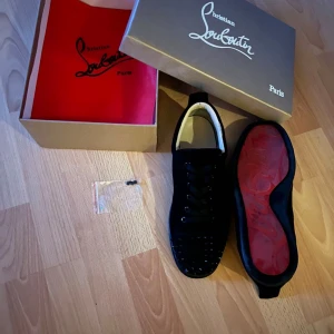 Svarta sneakers med nitar från Christian Louboutin - Säljer ett par svarta sneakers från Christian Louboutin med svarta nitar på tån och klassisk röd sula. Skorna har snörning och är tillverkade i skinn med en stilren och lyxig design. Kommer med originalkartong och dustbag. Skriv för mera info, priset är inte hugget i sten!😁Skorna har en enstaka defekt där av priset