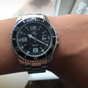 Silverfärgad Zhowe klocka med svart urtavla - Helt oanvänd stilren klocka från Zhowe med silverfärgad länk i metall och svart urtavla. Klockan har tydliga siffror, datumvisning och vridbar ring runt urtavlan. Perfekt accessoar för dig som gillar klassisk design med sportig känsla.