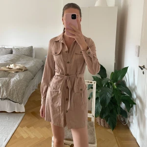 Beige skjortklänning med knappar och bälte - Säljer en beige skjortklänning med långa ärmar, knappar framtill och knytbälte i midjan. Klänningen har två stora fickor framtill och klassisk krage. Perfekt för en stilren och avslappnad look.