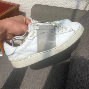Vita sneakers Valentino Garavani - Säljer ett par vita sneakers från Valentino Garavani med grå detalj över mitten och klassisk snörning. Skorna har en platt sula och rund tå, samt diskreta nitar baktill på sulan. Perfekta för dig som gillar stilrena och exklusiva sneakers.