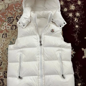 Vit dunväst från Moncler med huva - Säljer en snygg vit dunväst från Moncler med avtagbar huva och två dragkedjefickor framtill. Västen har en hög krage och klassisk Moncler-logga på bröstet. Perfekt för lager-på-lager under kyliga dagar. Tags samt påse medföljs.