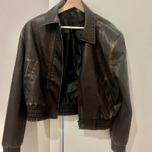 Brun skinnjacka - Brun skinnjacka med krage och resår vid midja och ärmslut. Jackan är i kort modell och axlarna är vadderade. En sliten look.