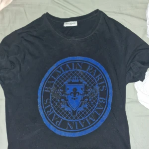 Svart t-shirt med blått tryck från Balmain - Säljer en svart t-shirt från Balmain Paris med stort blått tryck framtill. T-shirten har rund halsringning och klassisk passform. Perfekt för dig som gillar exklusiva märken och snygga detaljer.