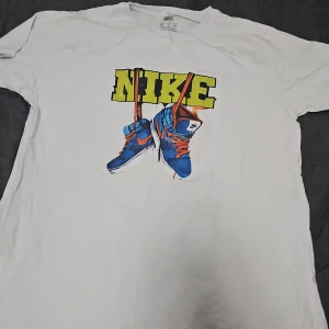 Vit t-shirt från Nike med sneakers-tryck - Vit t-shirt från Nike med stort färgglatt tryck framtill som visar blå sneakers och texten NIKE i gult. Klassisk passform och korta ärmar. Perfekt för en sportig och avslappnad stil.