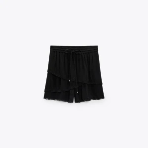 Svarta volangshorts från Zara - Svarta shorts från Zara med volangdetaljer och små bollar längs kanterna. De har snörning i midjan och är tillverkade i ett lätt, luftigt tyg som passar perfekt för varma dagar. Snygg och trendig modell med avslappnad passform. Aldrig använd och prislapp kvar!