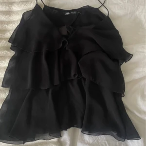 Svart volangig linnetopp från Zara - Supersnygg svart linnetopp från Zara med tunna axelband och flera lager volanger. Toppen har knytning framtill och är tillverkad i ett lätt, transparent tyg som ger en luftig känsla. Perfekt för dig som gillar en flowy och trendig look nu till sommaren, har använts ett fåtal gånger och visar inga tecken på användning 