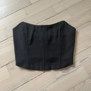 Svart korsett-topp - Säljer en snygg svart korsett-topp med markerade sömmar framtill som ger en stilren och edgy look. 