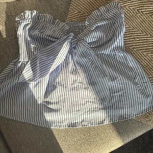Blåvit randig axelbandslös topp - 36/38 står 38 men jag upplever😂.. Superfin axelbandslös topp med blå och vita ränder och stor knytrosett framtill. Volangkant upptill och smockad rygg ger en söt och somrig vibe. Perfekt till jeans eller kjol för en fräsch look.