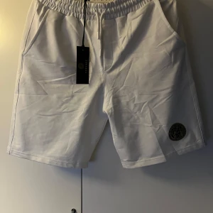 Vita Versace shorts med snörning - Snygga vita shorts från Versace med elastisk midja och snörning. Shortsen har två sidofickor och en ikonisk svart och guld Versace-logga broderad på ena benet. Perfekta för sommaren och ger en clean och lyxig vibe.