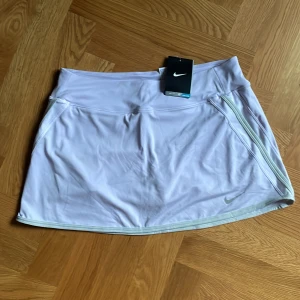 Nya Ljuslila sportkjol med innerbyxa från Nike - Sportig ljuslila kjol från Nike med inbyggda shorts och Dri-FIT-teknologi. Kjolen har en bred midjeresår och diskret Nike-logga framtill. Perfekt för dig som gillar att röra på dig och vill ha en snygg och bekväm look. Helt nya med tag kvar