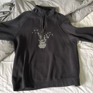Svart half zip hoodie med rhinestones - Svart hoodie med half zip och hög krage. På Tröjan har långa ärmar och är i mjukt bomullsmaterial. Perfekt för dig som gillar unika detaljer och comfy stil. 
