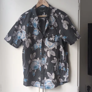 Retrofit blommig skjorta, storlek L - Snygg kortärmad skjorta från Retrofit med grå bas och stora blommor i blått och vitt. Skjortan har klassisk krage, knäppning framtill och en bröstficka. Perfekt för dig som vill sticka ut med ett coolt blommönster och skön passform.