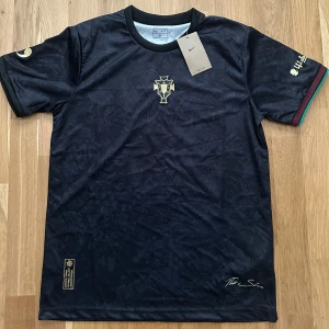 Svart Ronaldo fotbollströja  - Svart fotbollströja från 9commafootball x The Sin i storlek M. Tröjan har guldfärgade broderade detaljer, diskret mönster i tyget och kontrastfärgade muddar i vinrött och grönt. Tillverkad i lätt och ventilerande material, perfekt för match eller träning.