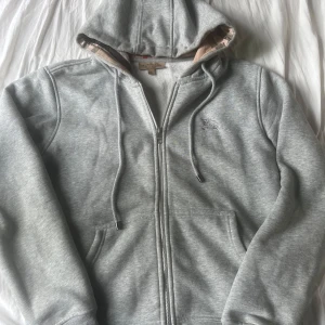 Burberry zip grå - Snygg grå hoodie från Burberry med klassiskt rutigt foder i huvan och broderad logga på bröstet. Tröjan har dragkedja framtill, snörning i huvan och två fickor. Perfekt för dig som gillar stilrena och exklusiva plagg. Köpt från plick, ny och ej defekter eller smuts. Pris kan diskuteras vid snabb affär