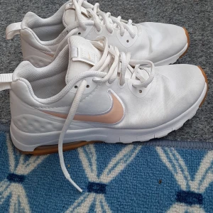 Vita Nike sneakers med persikofärgad swoosh - Snygga vita Nike sneakers med persikofärgad swoosh och gummisula i ljusbrun ton. Skorna har snörning och är tillverkade i syntetmaterial och textil. Perfekta för dig som gillar en clean och sportig look med en twist av färg.