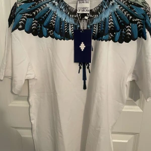 Vit t-shirt Marcello Burlon med blå fjädrar - Snygg vit t-shirt från Marcelo Burlon County of Milan med ikoniskt blått och svart fjädertryck runt halsen. Klassisk passform och korta ärmar, perfekt för dig som vill sticka ut med en unik design. Materialet är mjuk bomull för maximal komfort.